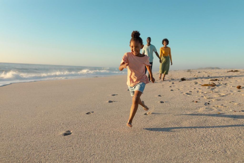 A photo of a girl running on the beach with her parents behind here watching
